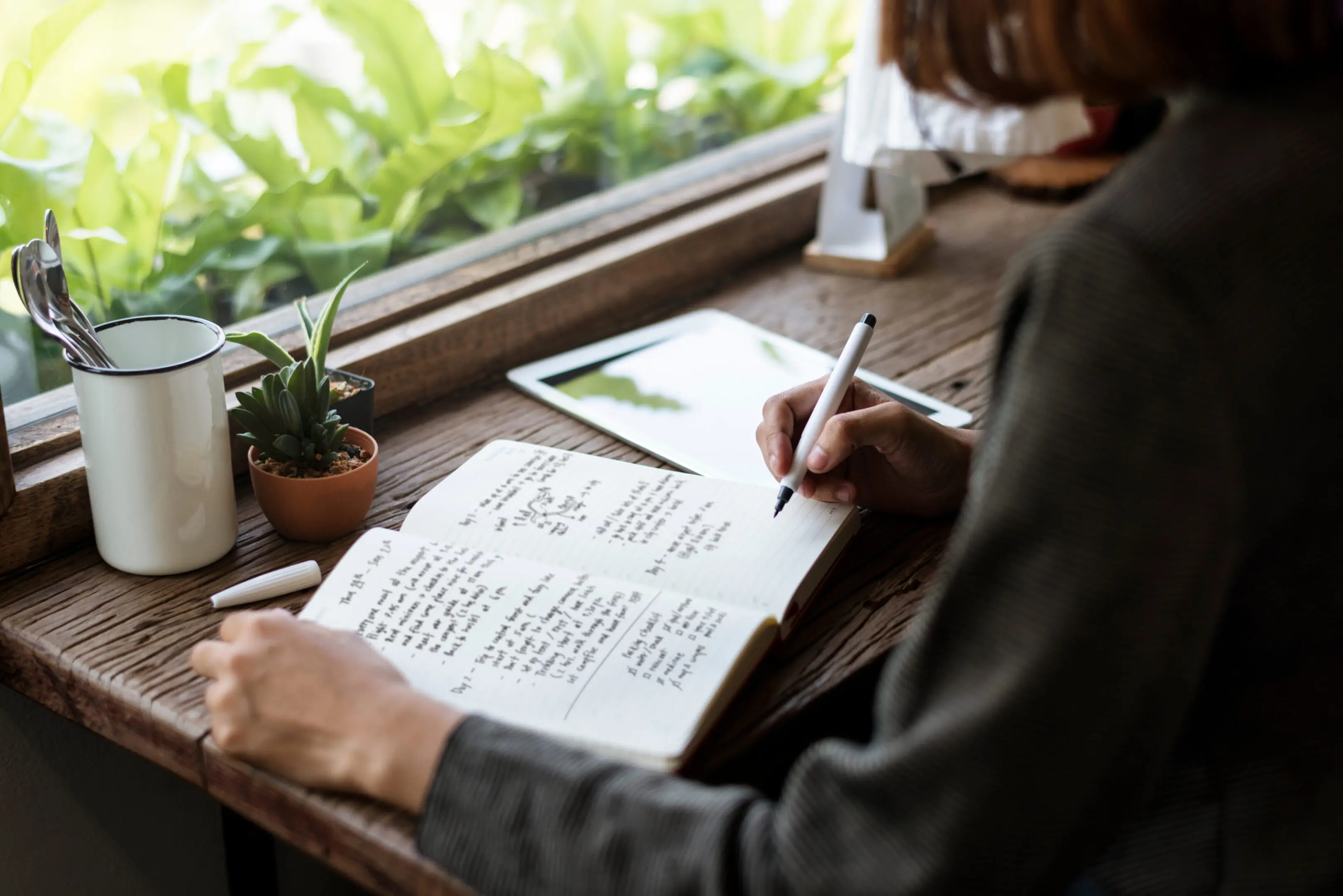 Person writing notes in a notebook by the window with plants nearby.